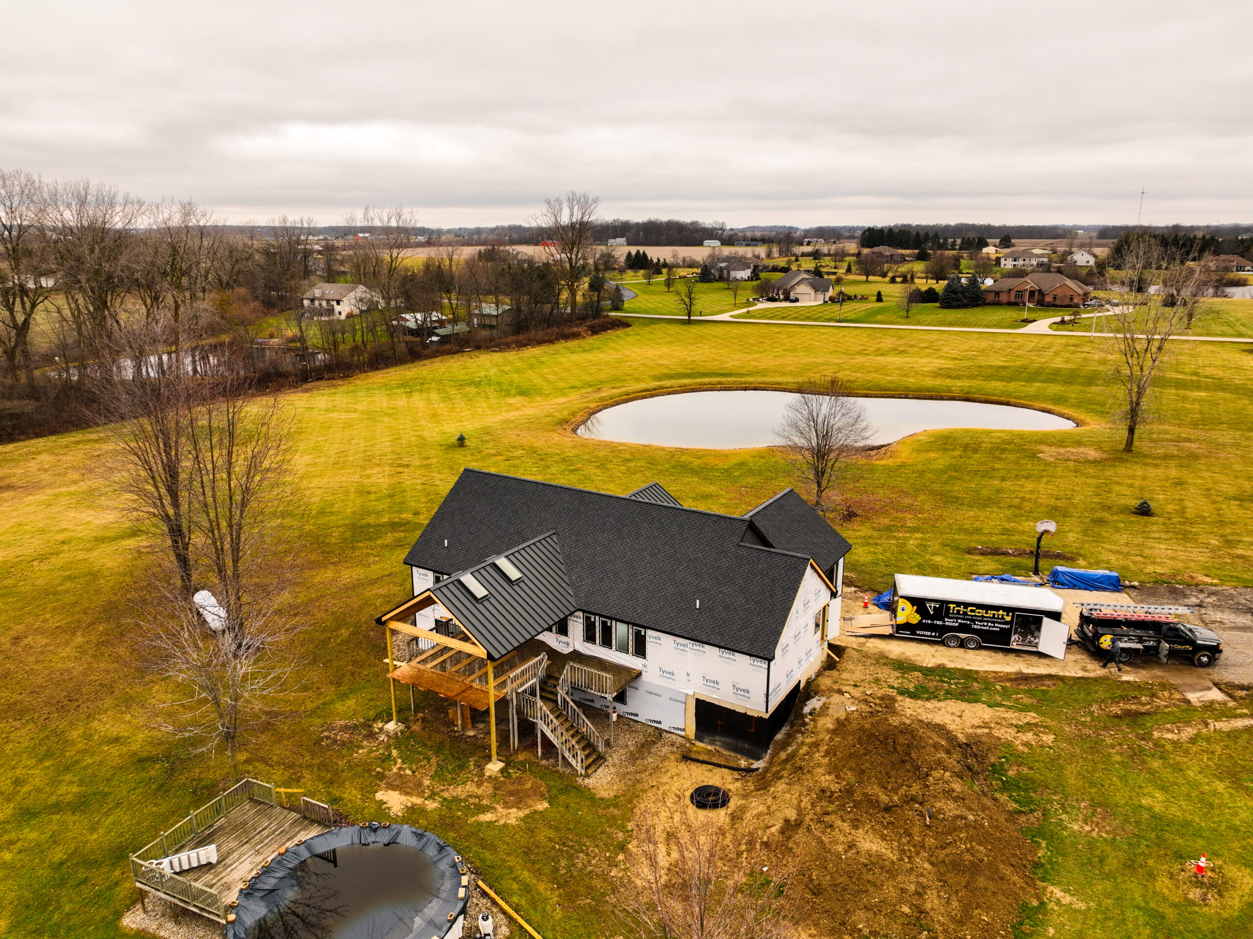 Aerial view of a house under construction with a dark shingle roof, metal porch roof, and Tri-County vehicles nearby