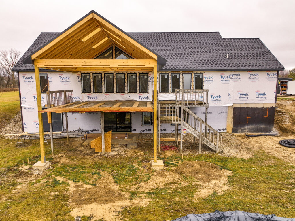 Rear view of a house under construction with a shingle roof and unfinished timber-framed covered porch