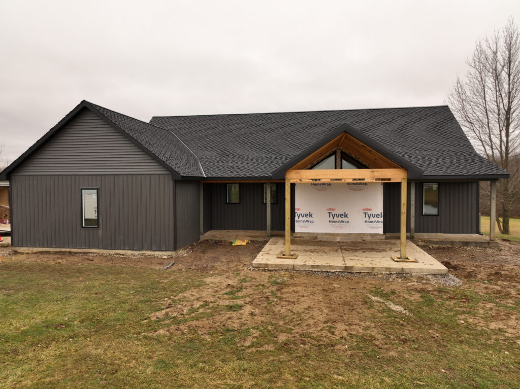 House under construction with a dark shingle roof, gray siding, and an unfinished timber porch